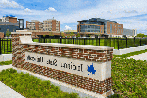 A large brick building with large windows is visible in the background of the image on the left, with three campus towers in the distance on the far right of the frame. In the foreground is a brick campus entryway surrounded by green grass, with a blue Sycamore leaf logo and “Indiana State University” on the sign.